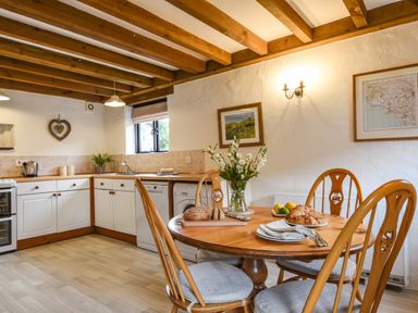 A kitchen with wooden cabinets a round wooden dining table with chairs and flowers at Bramble Cottage in Ringmore