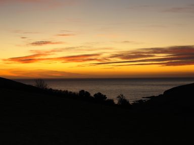 A coastal landscape at sunset with a dark hillside and trees in the foreground and the sea in the background at Bramble Cottage in Ringmore