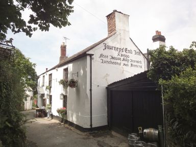A white building with hanging flower baskets and barrels outside on a narrow road at Journeys End Inn