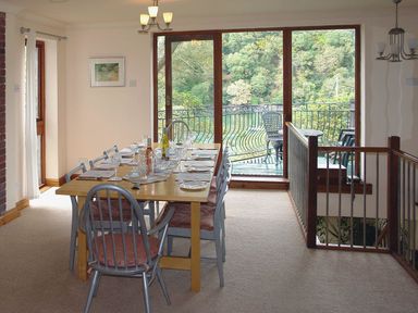 A dining room with a wooden table set with glasses and bottles next to large windows overlooking a balcony with chairs and trees at Galanda Treetops in Newton Ferrers