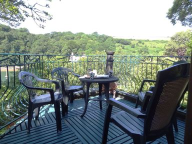 A balcony with plastic chairs and a round table overlooking trees and a river at Galanda Treetops Newton Ferrers
