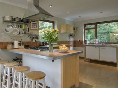 A kitchen with a central island with stools pots hanging on wall and large windows at Galanda Treetops in Newton Ferrers