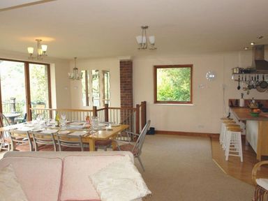 An open plan dining and kitchen area with a wooden dining table and chairs a kitchen island with stools and a window showing greenery outside at Galanda Treetops in Newton Ferrers