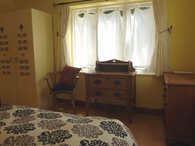 A bedroom with patterned bedspread wooden chair with cushions wooden drawers and white curtains at Galanda Treetops in Newton Ferrers