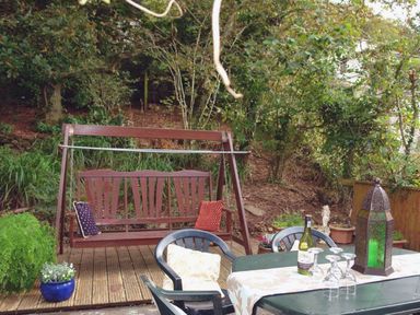 An outdoor wooden swing with two cushions and a table with chairs set on a deck surrounded by plants and trees
