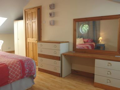A bedroom with a bed red patterned bedspread a wooden door a chest of drawers a dressing table with a mirror and a window reflection at Galanda Treetops in Newton Ferrers
