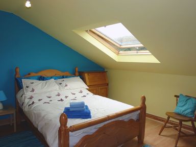 A bedroom with a wooden bed with butterfly bedding and towels on it a skylight window a wooden chair with blue cushions and a small wooden table with a blue lamp at Galanda Treetops in Newton Ferrers