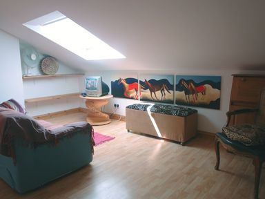 An attic room with a small TV on a wooden spiral table a sofa a long beige ottoman a chair and a triptych painting of horses on the wall at Galanda Treetops in Newton Ferrers