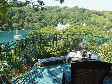 A balcony with two chairs and a table overlooking a river with a boat and trees at Galanda Treetops in Newton Ferrers