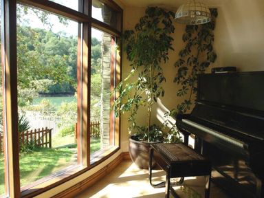 A room with a black piano and a stool next to a large window overlooking a garden and trees at Galanda Treetops in Newton Ferrers