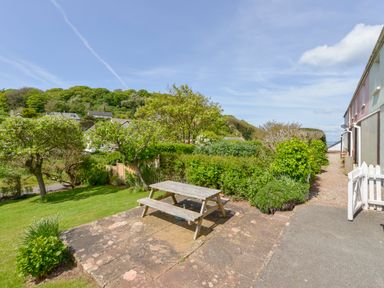 A garden area with a wooden picnic table on a stone patio surrounded by trees and bushes at 4 Coastguards Cottage in Hope Cove