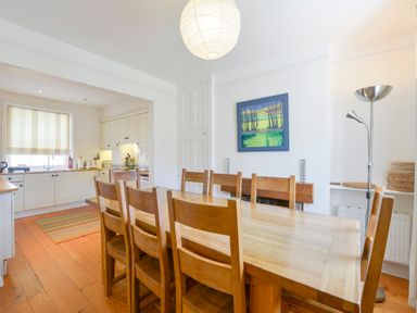 A dining room with a wooden table and chairs next to a kitchen with white cabinets and a window at 4 Coastguards Cottage in Hope Cove
