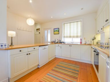 A kitchen with white cabinets wooden countertops a striped rug and a window with a blind at 4 Coastguards Cottage in Hope Cove
