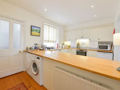 A kitchen with white cabinets a wooden countertop washing machine and dryer under the counter and a striped window blind at 4 Coastguards Cottage Hope Cove