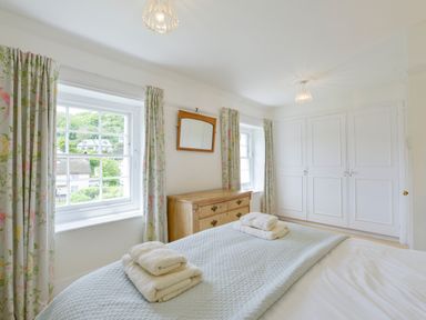 A bedroom with a bed folded towels on a blanket windows with floral curtains a wooden dresser and white wardrobes at 4 Coastguards Cottage in Hope Cove