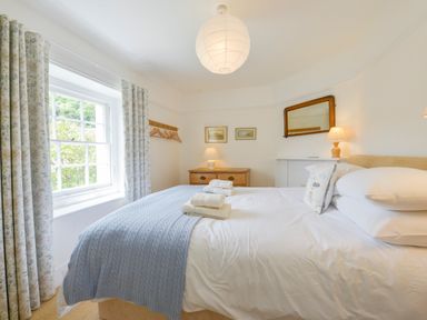 A bedroom with a bed pillows folded towels bedside tables lamps a window with curtains and framed pictures on the wall at 4 Coastguards Cottage in Hope Cove