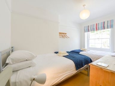 A bedroom with two single beds with white bedding and blue throws a wooden desk a window with striped blinds and a wall rack with hangers at 4 Coastguards Cottage in Hope Cove