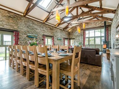 A dining area with a long wooden table and chairs beside a sitting area with brown sofas and a fireplace at Mill Barn in Woolston near Salcombe
