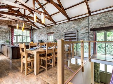 A dining area with wooden table and chairs next to a living room with leather sofas in a room with exposed stone walls and wooden beams at Mill Barn in Woolston near Salcombe