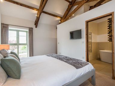 A bedroom with a bed and pillows a window with curtains a wall-mounted television and an ensuite bathroom with a bathtub at Mill Barn in Woolston near Salcombe