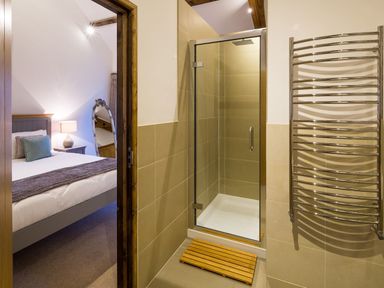 A bathroom with a glass shower enclosure a wooden mat and a chrome towel rack next to a bedroom with a bed a side table and a mirror at Mill Barn in Woolston near Salcombe