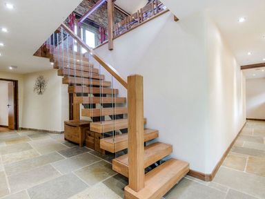 A hallway with stone tile flooring and a wooden staircase with glass panels at Mill Barn in Woolston near Salcombe