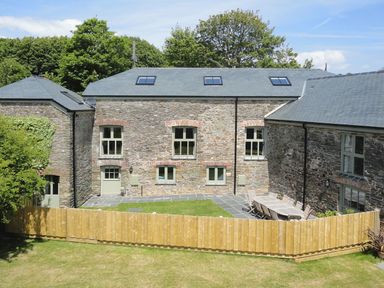 An outdoor area with stone buildings trees and a wooden fence surrounding a patio with a table and chairs at Mill Barn in Woolston near Salcombe