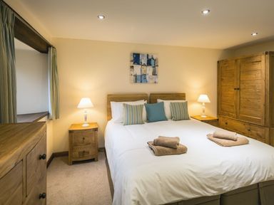 A bedroom with a double bed pillows bedside tables lamps wooden wardrobe and chest of drawers at Mill Barn in Woolston near Salcombe