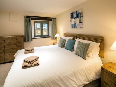 A bedroom with a double bed with pillows and folded towels a wooden dresser two bedside tables with lamps a window with curtains and a painting on the wall at Mill Barn in Woolston near Salcombe