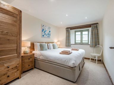 A bedroom with a double bed bedside tables lamps a wooden wardrobe a window with curtains and a chair at Mill Barn in Woolston near Salcombe