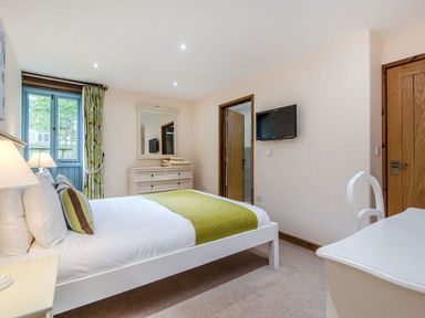 A bedroom with a white bed and green bed runner a white dresser with a mirror a wall mounted tv and wooden doors at Mill Barn in Woolston near Salcombe