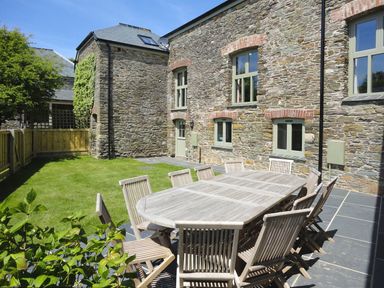 A stone building with multiple windows a wooden dining table and chairs on a tiled patio and a lawn enclosed by a wooden fence at Mill Barn in Woolston near Salcombe
