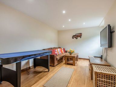 A living room with a ping pong table a brown sofa with cushions a wooden coffee table a flower vase a wall-mounted TV and wooden furniture at Mill Barn in Woolston near Salcombe