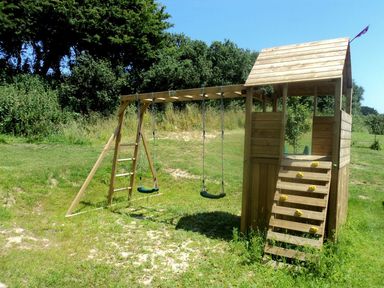 A wooden playhouse with swings and a climbing wall in a grassy outdoor area surrounded by trees at Mill Barn in Woolston near Salcombe
