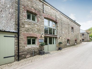 The exterior of a stone barn with green doors and windows and a sign that reads Mill Barn in Woolston near Salcombe