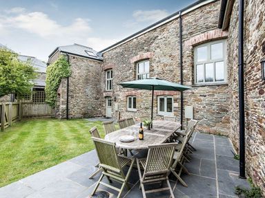 An outdoor patio with a wooden dining table and chairs under an umbrella next to a stone building at Mill Barn in Woolston near Salcombe