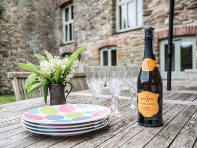 An outdoor wooden table with a bottle of prosecco four empty glasses a small bouquet of flowers and a stack of colorful plates at Mill Barn in Woolston near Salcombe