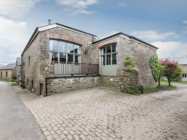 A stone building with large windows and a wooden railing on a cobblestone driveway with trees nearby at Mill Barn in Woolston near Salcombe