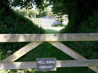 A wooden gate with a sign saying mill barn orchard and a path leading to a shed surrounded by greenery at Mill Barn in Woolston near Salcombe