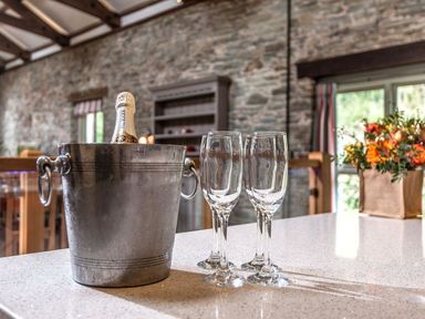 A bottle of champagne in an ice bucket with four empty champagne flutes on a countertop in a room with stone walls and a window with flowers nearby at Mill Barn in Woolston near Salcombe