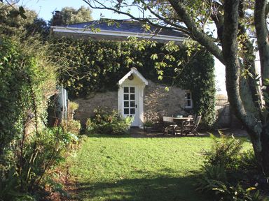 A garden with grass trees and outdoor furniture in front of a stone house covered with ivy at Coach House Cottage in Strete near Dartmouth