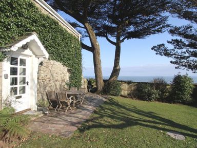 An outdoor patio area with a wooden table and chairs next to a stone house covered in ivy and a view of the sea at Coach House Cottage in Strete near Dartmouth