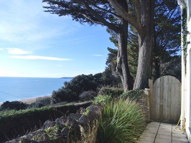 A stone wall and tall trees next to a wooden gate with a view of the sea at Coach House Cottage in Strete near Dartmouth