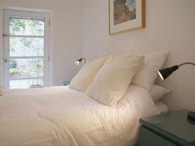A bedroom with a bed white pillows bedside tables with lamps and a glass door showing outdoor plants at The Gallery in Aveton Gifford