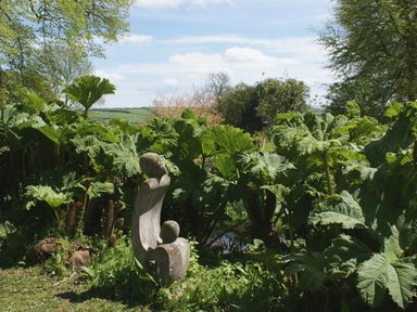A garden with a stone sculpture surrounded by large green leafy plants and trees in the background at The Gallery in Aveton Gifford