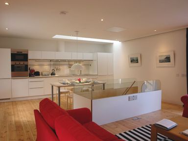 A modern kitchen with white cabinets and a dining table next to a living area with red sofas and wooden flooring at The Gallery in Aveton Gifford