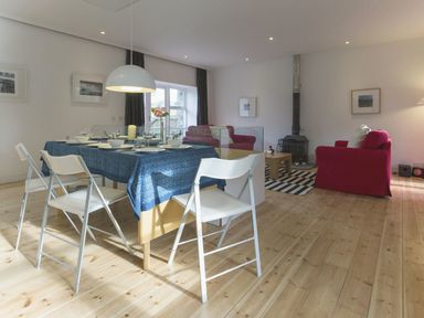 A dining table with white chairs and blue tablecloth next to a sitting area with red sofas and a wood stove at The Gallery in Aveton Gifford