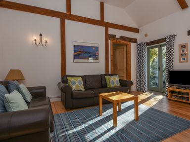 A living room with two leather sofas a wooden coffee table a rug wall beams framed artwork double doors and a television on a stand at Bradbridge Barn in Newton Ferrers