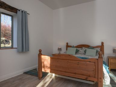 A bedroom with a wooden bed frame, bedside tables with lamps, a window with blue curtains, and a striped rug on the floor at Bradbridge Barn in Newton Ferrers