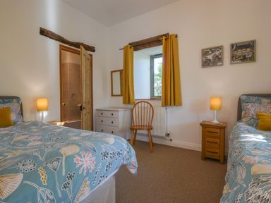 A bedroom with two beds bedside tables with lamps a wooden chair a window with yellow curtains and a door to a bathroom at Bradbridge Barn in Newton Ferrers
